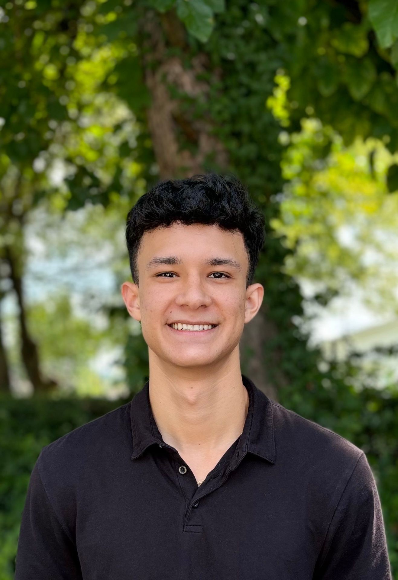 Young man smiling in a black shirt with green foliage background.