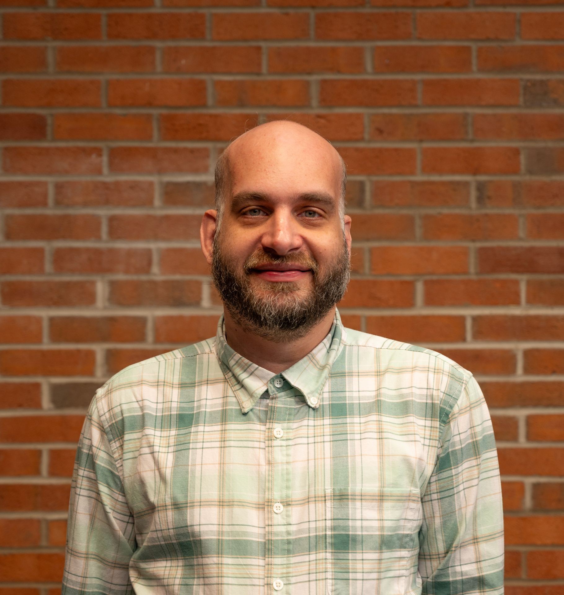 Person wearing a green and white plaid button-up shirt standing in front of a brick wall.