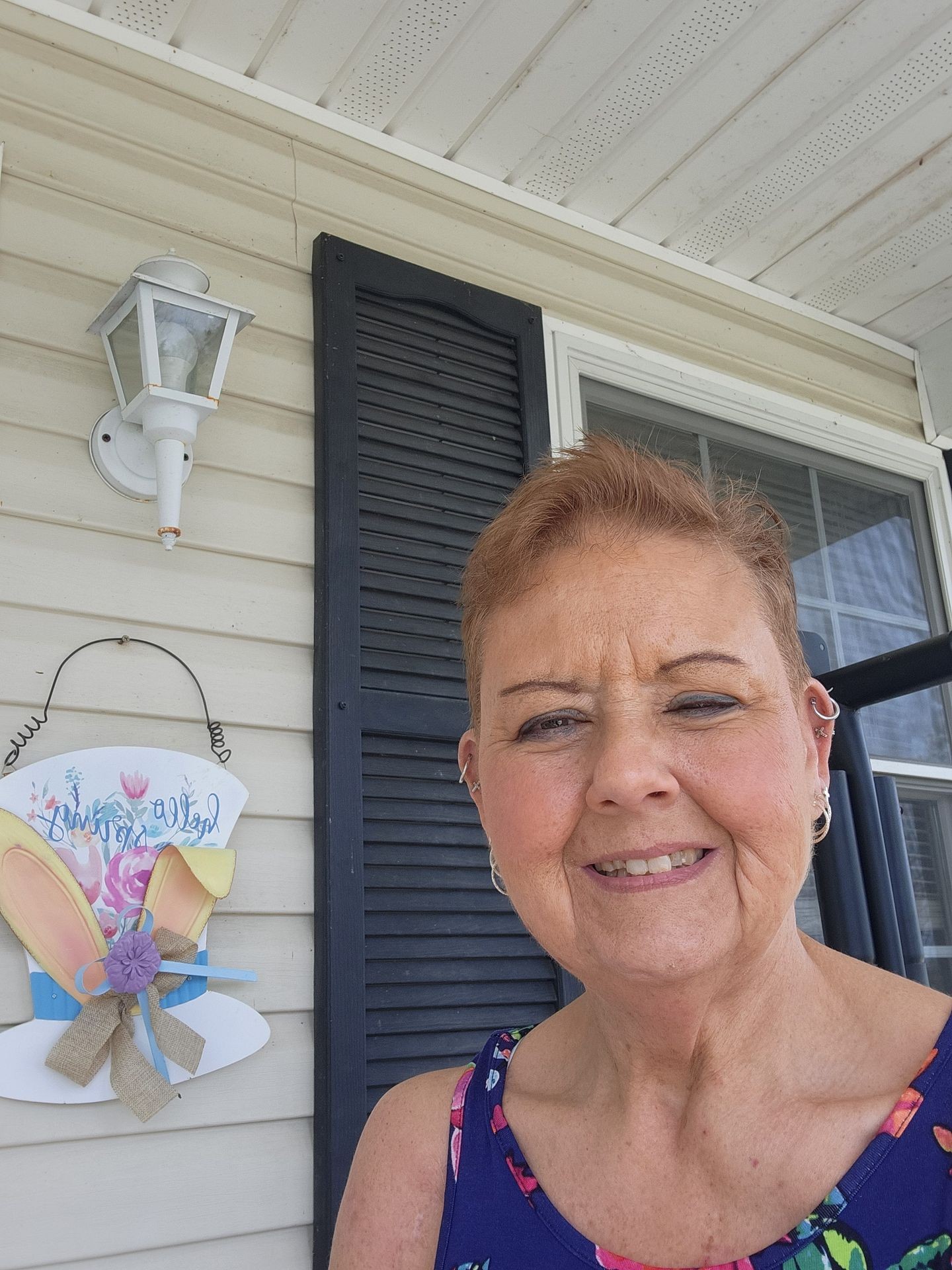 Person wearing a colorful floral dress standing on a porch with a wall-mounted light and decorative sign.