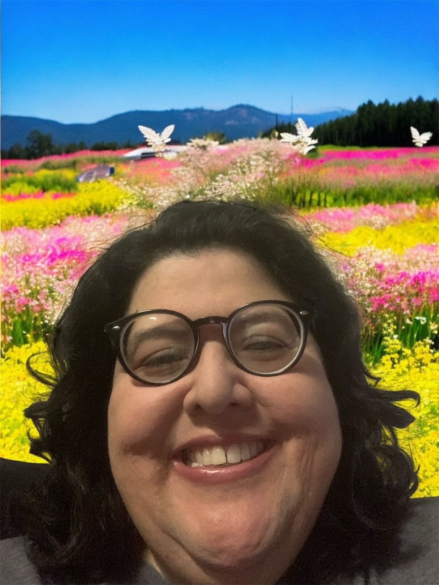 Person smiling in front of a vibrant field of flowers with mountains and blue sky in the background.