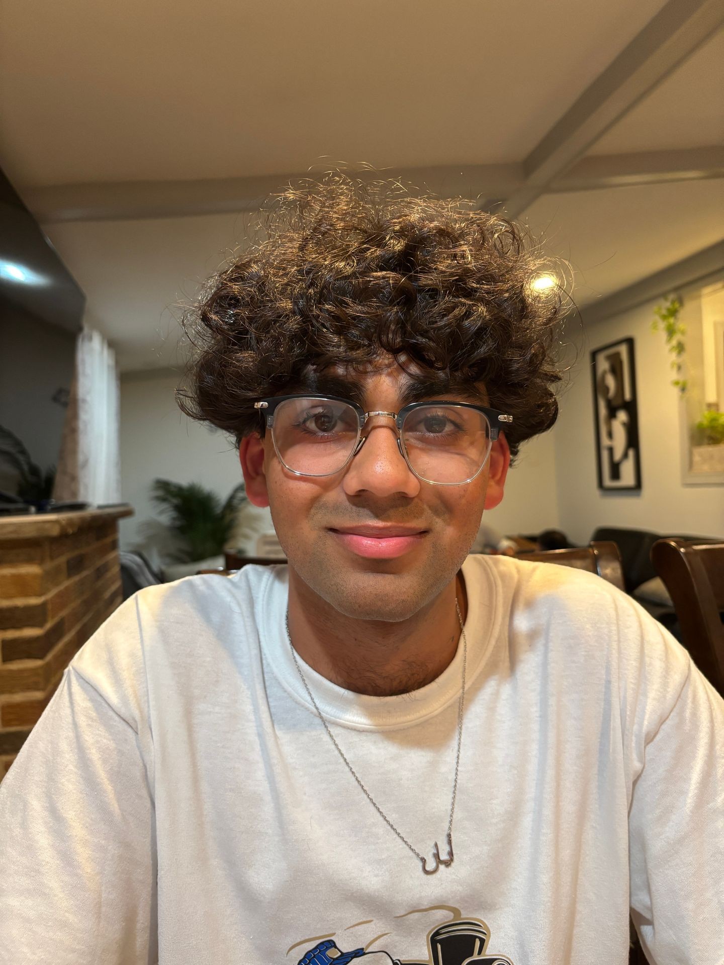 Person with curly hair and glasses smiling, wearing a white shirt indoors with a brick background.