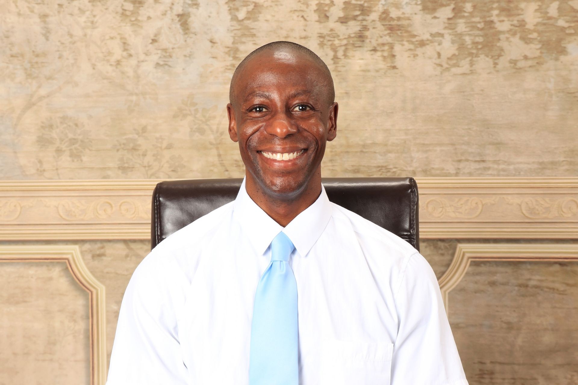 Smiling man in a white shirt and blue tie seated against a vintage-style decorative background.