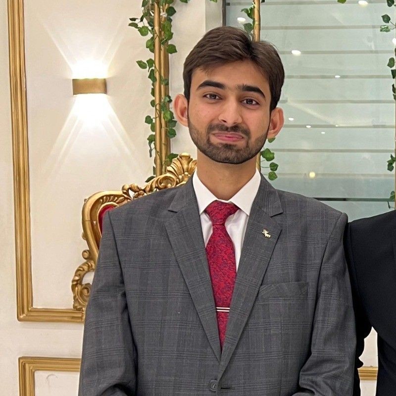 Man in a grey suit and red tie standing indoors with decorative wall and plants.