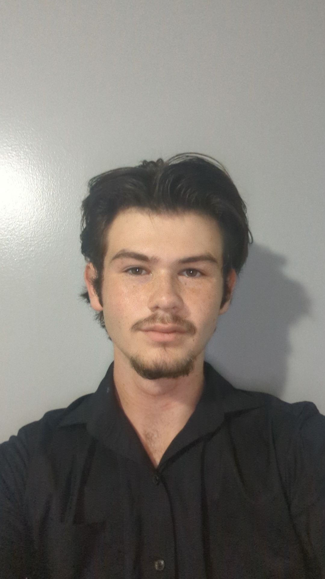 Person with short dark hair and facial hair wearing a black shirt against a plain gray background.